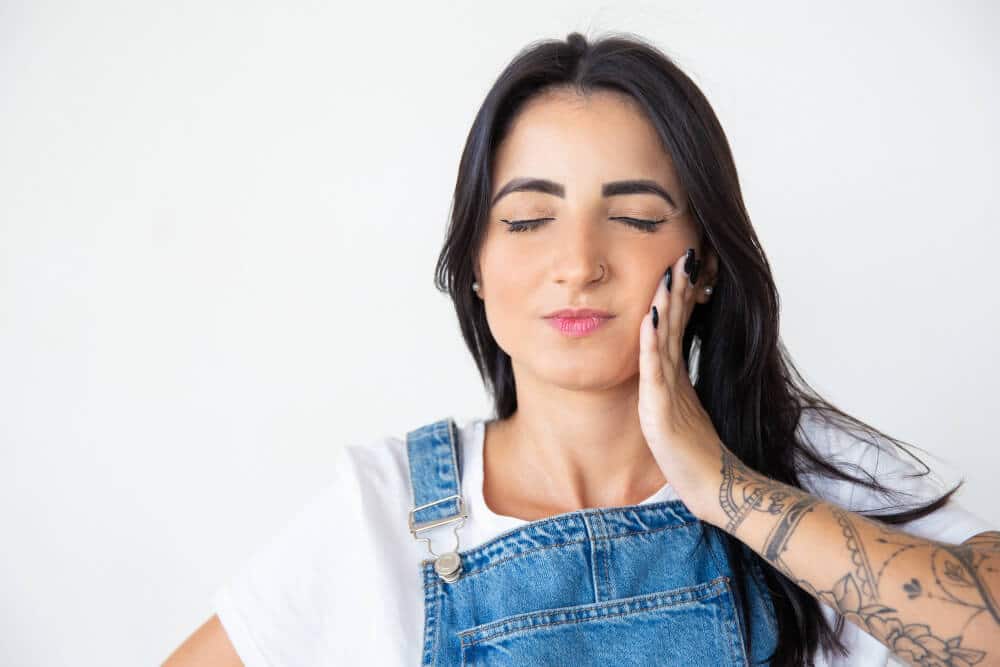 Moça jovem com a mão esquerda na região da mandíbula e cara de dor, representando algumas dores na Articulação Temporomandibular